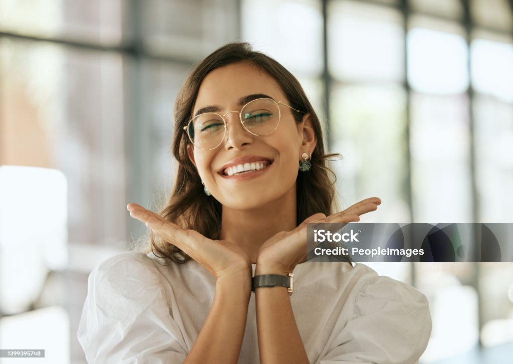 Cheerful business woman with glasses posing with her hands under her face showing her smile in an office. Playful hispanic female entrepreneur looking happy and excited at workplace Happiness Stock Photo