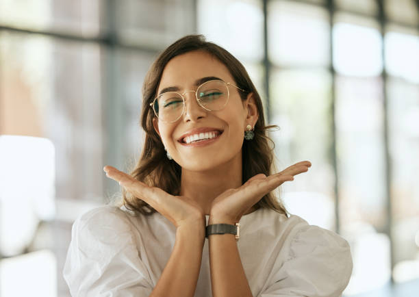 wesoła kobieta biznesu w okularach pozująca z rękami pod twarzą pokazująca swój uśmiech w biurze. zabawna latynoska przedsiębiorcza kobieta wyglądająca na szczęśliwą i podekscytowaną w miejscu pracy - smiling zdjęcia i obrazy z banku zdjęć