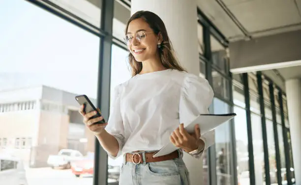 Photo of Happy business woman texting on smartphone holding file and walking in modern office. Smiling female entrepreneur using mobile app or browsing social media