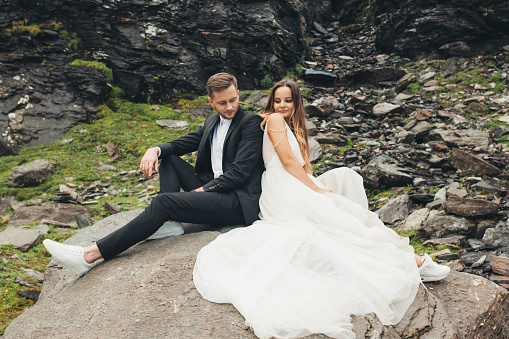 Portrait of a couple taking a photo shoot on their wedding day sitting on a stone back to back on the background of a rocky mountain hill. Beautiful portrait. Wedding travel. Honeymoon trip.