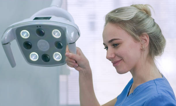 pretty female nurse adjusting dental light in clinic. dentist assistant prepares lighting equipment before treatment - dentist office nurse doctors office blond hair imagens e fotografias de stock