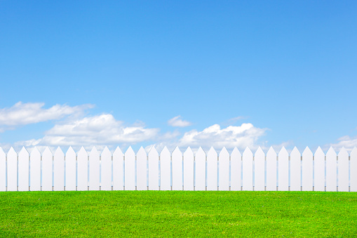 A white picket fence sits on a brilliant green lawn in front of a blue sky with clouds that hug the horizon in the distance.