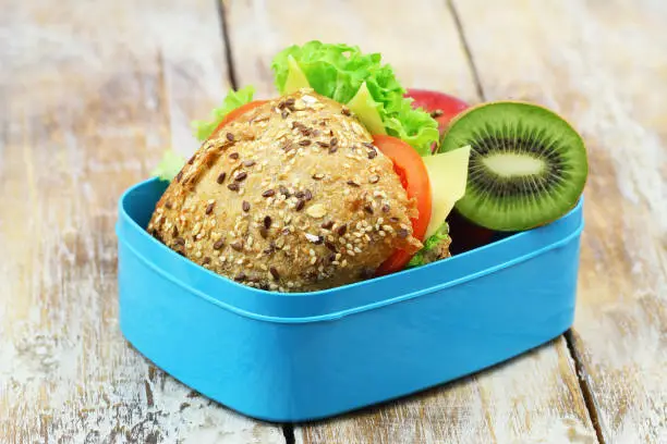 Healthy lunchbox containing whole grain cheese roll with lettuce and tomato and kiwi fruit