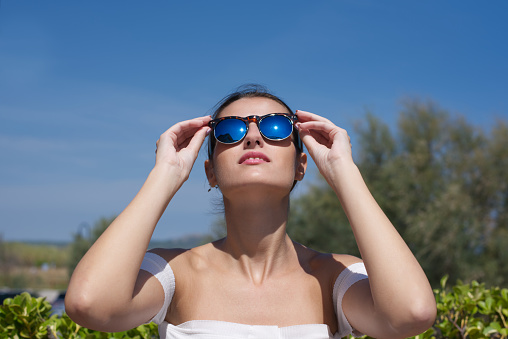 Portrait of young beautiful fashionable woman with sunglasses looking up. Woman enjoying a sunny day on a terrace facing the sea in summer.