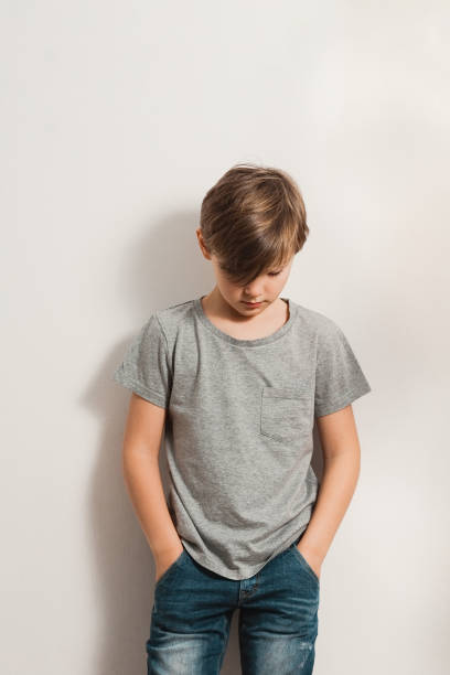 cute sad boy bowing his head down, leaning to white wall a cute boy stands next to white wall, grey t-shirt, blue jeans, hands in pockets, bowing his head down sad child standing stock pictures, royalty-free photos & images