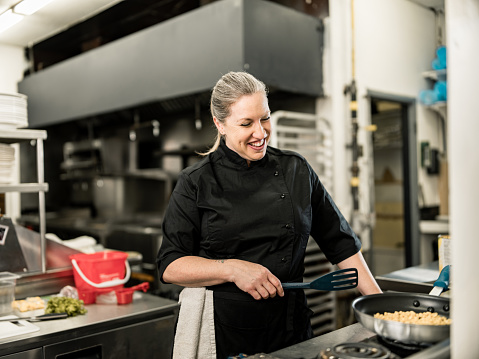 Portrait of female chef at the commercial kitchen. Mature Caucasian woman dressed in black chef's uniform. Interior of hotel commercial kitchen during day.
