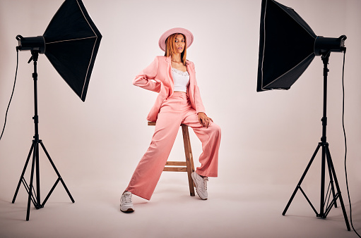 Portrait of a young mixed race female posing in trendy fashionable clothing while chilling on a chair in a studio shoot. Hispanic woman showing the latest fashion collection with a cool style and hat