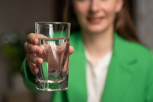 Woman stretches out hand holding glass with clean water to drink on blurred background. Young lady in green jacket keeps healthy lifestyle closeup