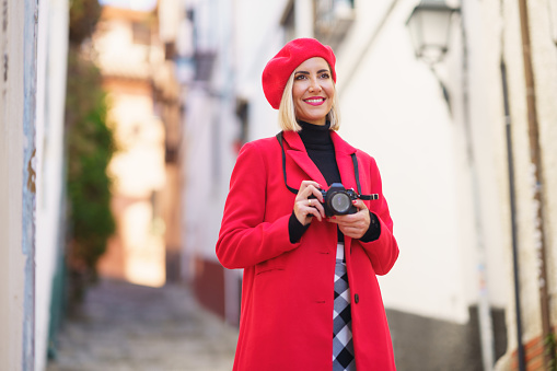Cheerful young female traveler in fashionable red coat and beret, smiling and looking away, while standing on narrow street between residential buildings and taking photos on camera