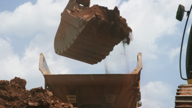 Slow Motion Shot of a Bucket of an Excavator Digging, Picking Up Dirt, and Dumping It into the Back of a Dump Truck at a Sunny Construction Site