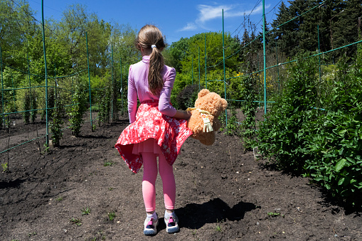 A 7 year old girl with dark hair in a pink suit stands alone in a green garden on the ground with a brown bear