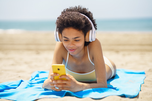 African American woman lying on a towel on the sand on the beach using her smartphone to check Social Media. African American girl tanning.