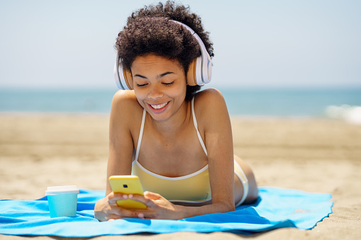 Smiling black woman lying on a towel on the sand on the beach using her smartphone to check Social Media. African American girl tanning.