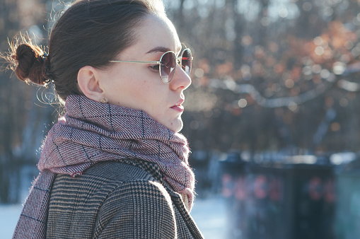 Street style fashion portrait charming young woman in trendy casual clothes, side view. Brown overcoat, light red scarf, sunglasses. Winter collection natural look