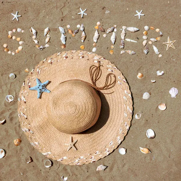 Straw hat on beach sand beside word Summer made of seashells