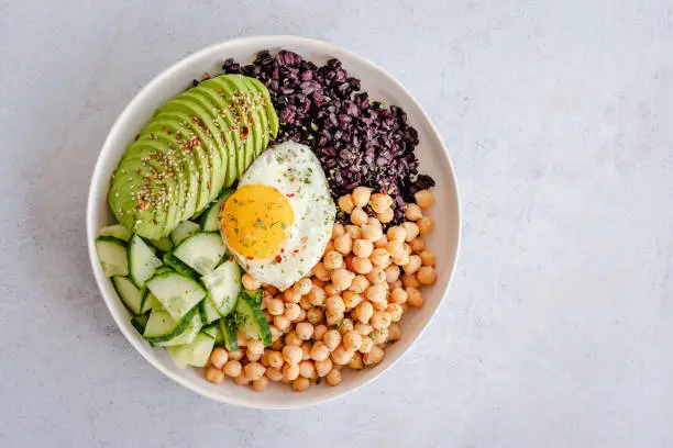 Overhead view of a bowl with chickpeas, black rice, avocado, egg and cucumber on a minimal background with copy space on the right side