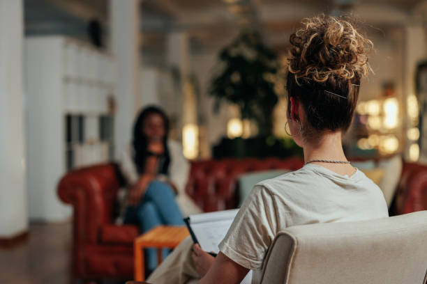 View from behind of female psychotherapist on session Focus on female psychiatrist writing information of african female patient while sitting on sofa and talking about his illness mental health professional stock pictures, royalty-free photos & images