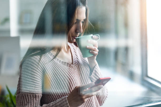 istock Pretty young woman using her mobile phone while drinking coffee near to the window in the living room at home 1399433182
