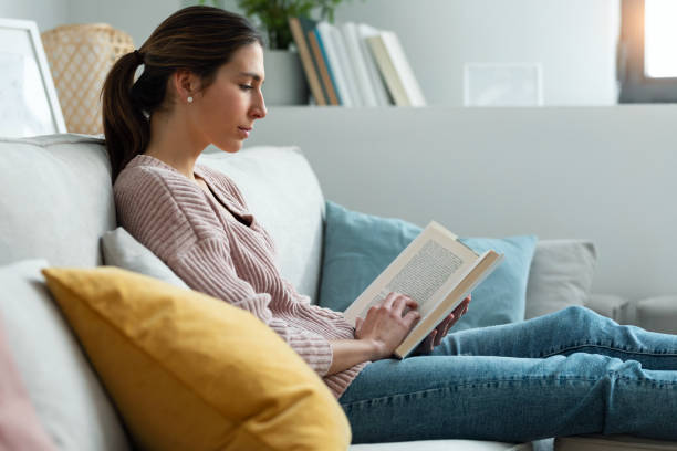 istock Pretty young woman reading a book while sitting on sofa at home. 1399433286