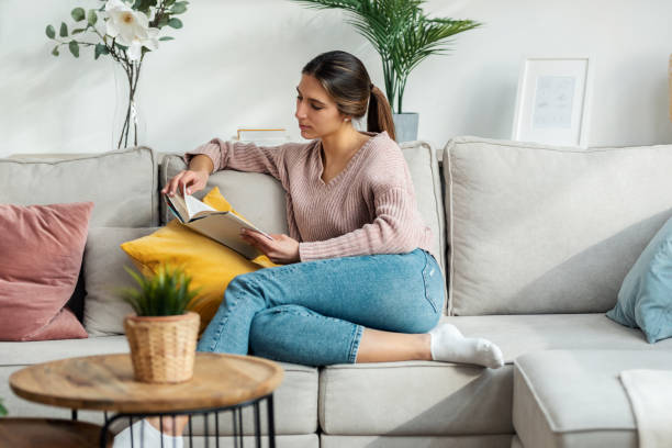 istock Pretty young woman reading a book while sitting on sofa at home. 1399433466