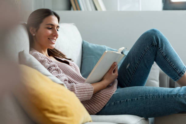 istock Pretty young woman reading a book while sitting on sofa at home. 1399433533