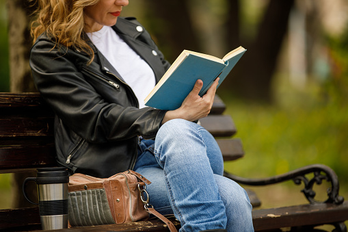 Cut out shot of cool mid adult blond sitting on a bench at the park, relaxing, enjoying coffee from a travel mug and reading a book.