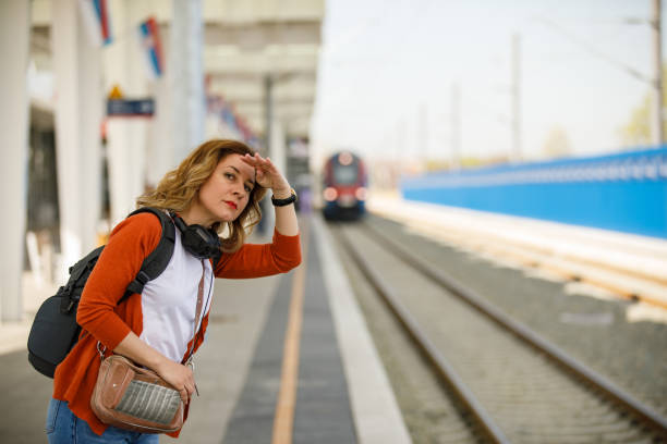 Selective focus shot of mid adult woman going on a trip. She is standing on a railroad station platform, covering her eyes from the sun and looking in the distance while waiting for her train to arrive.