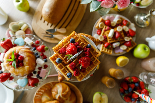 Selective focus shot of tea stand with delicious waffles decorated with fresh berries, a cake on a cake stand and more waffles served on a plate on a dining table, among other delicious sides for a tasty breakfast, brunch or afternoon tea.