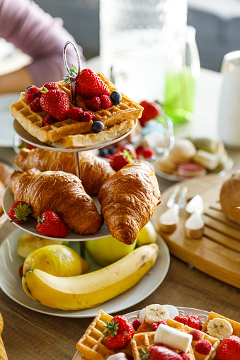 Close up shot of a tea stand with delicious waffles, croissants, banana, lemons and fresh berries served on a dining table to be enjoyed during a meal.