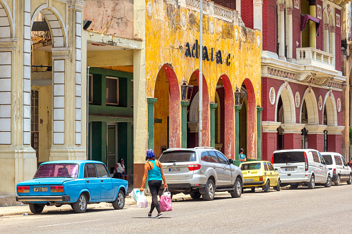 Havana, Cuba - May 18, 2022: A Cuban woman carries three bags as she crosses the street by the Cine Actualidades. Diverse cars are parked in the area. The old movie theater is a local landmark