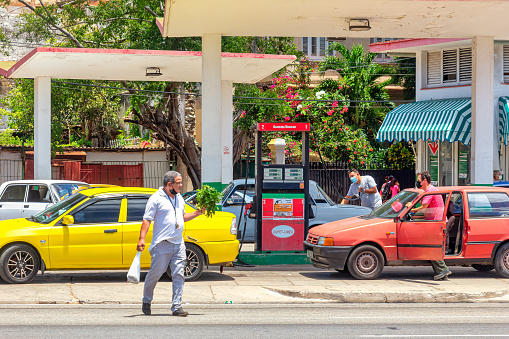 Havana, Cuba - May 18, 2022: A man with a bunch of vegetables and a bag crosses the street by a Cupet-Cimex gas station. Diverse cars are buying gasoline in the government-run business. Other incidental people are seen in the scene.