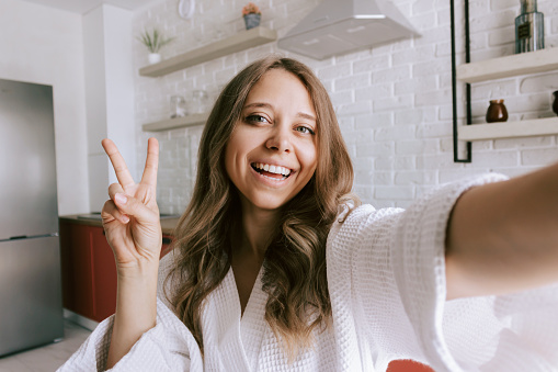 A young beautiful caucasian smiling blonde woman in a white robe takes a selfie showing a peace gesture with her v-shaped hand in the morning on background of the kitchen