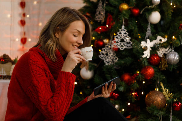 istock Young woman using smart phone and sipping coffee by the Christmas tree 1399443338