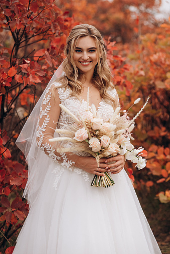 Bride holding boho bouquet against of trees with red leaves in autumn