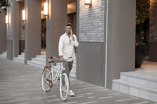 Young stylish man walks with bicycle around the city using mobile smartphone. Online communication. 5G internet connecting. Freelance lifestyle. Top view of modern pavement road for bike transport