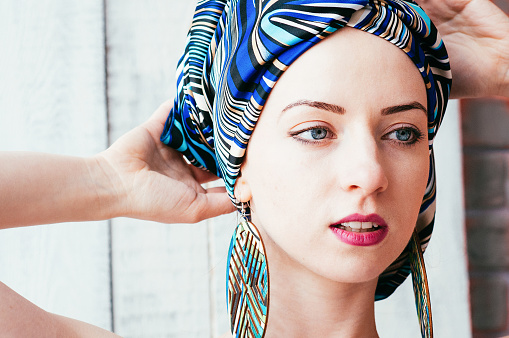 a close-up portrait of a pretty young woman wearing a colorful blue headscarf in an african manner, big round earrings, blue eyes, light background