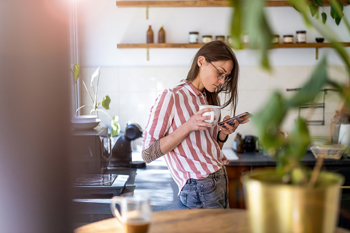 Young woman using smartphone while standing in her kitchen at home