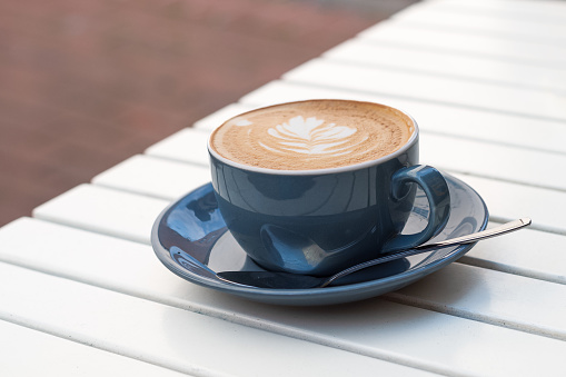 Shallow focus on the froth of flat white coffee in a blue cup and saucer with a teaspoon on the saucer. The coffee is on a white wooden slated table outside.