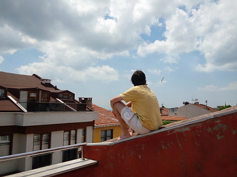 young man sitting on the roof of a house enjoying the view of the old city, seagulls flying in the cloudy sky on a summer day