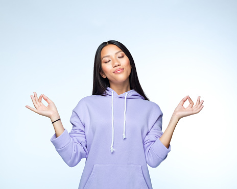 Portrait of young woman wearing lilac hoodie meditating with eyes closed and raised hands while standing against white background.