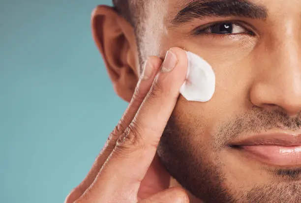 Photo of Closeup of one young indian man applying moisturiser lotion to his face while grooming against a blue studio background. Handsome guy using sunscreen with spf for uv protection. Rubbing facial cream on cheek for healthy complexion and clear skin