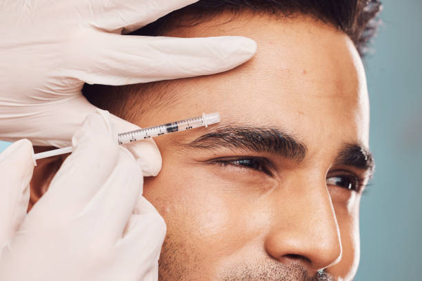 beau jeune homme indien recevant une injection de botox sur un fond de studio bleu. un gars métis reçoit un traitement pour réduire le vieillissement et combler les rides du visage - plastic protective glove wrinkle treatment medical instrument photos et images de collection