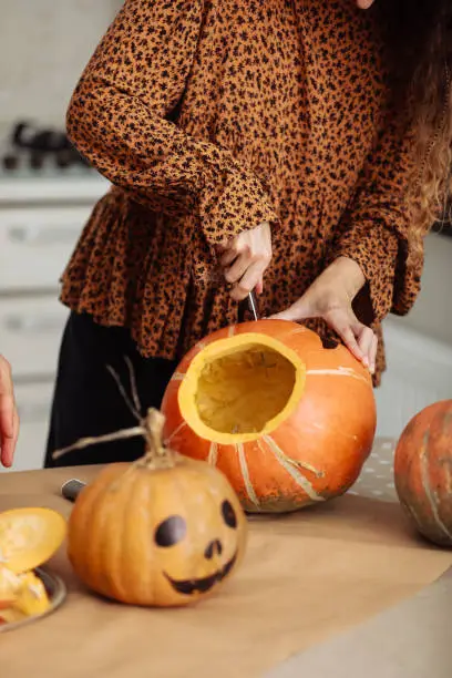 Photo of Close up of young woman carving Jack O Lattern from ripe orange pumpkin with knife on her wooden kitchen table. Female preparing all hallows eve Halloween party decorations. Background, copy space.