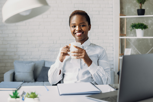Indoor portrait of pleased african american woman looking at camera while posing in office during coffee break. Businesswoman portrait. Stylish american black portrait. Business woman. Afro american girl.
