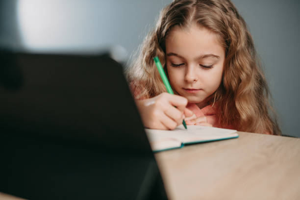 istock Caucasian little girl with tablet pc computer writing to notebook at home. Online education. Kid learn online learning school. Quarantine, pandemic. 1399469998