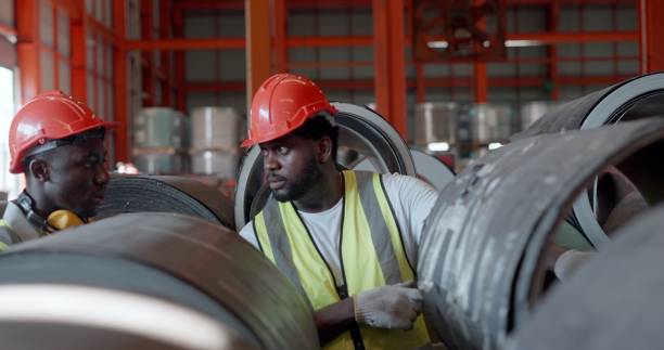 istock Team professional African American worker or foreman in metal work, wear personal protective equipment, talking and monitor roll steel the raw material store according to the list in paper, in production line at metal part manufacturing factory. 1399497453