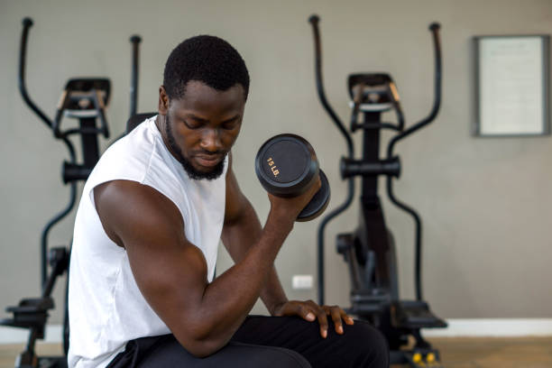 istock Young short curly black hair man with moustache and beard looking at his arm while working on his biceps, lifting barbell with one hand. There are cardio machines in the gym. 1399502137