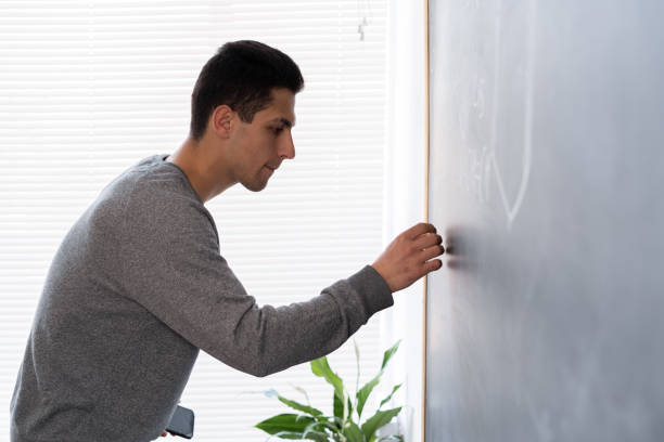 istock Young arab teacher leading lesson writes with chalk on blackboard, university student at chalk board, male arab 20s studying in class, side view tutor, education 1399502459