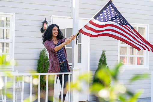 Mid adult woman hangs a american flag on her porch and celebrating Fourth of July. Independence day.