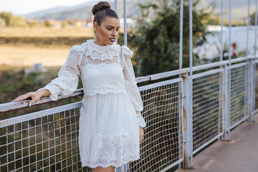 Beautiful young woman in white dress standing on foot bridge on sunny summer day